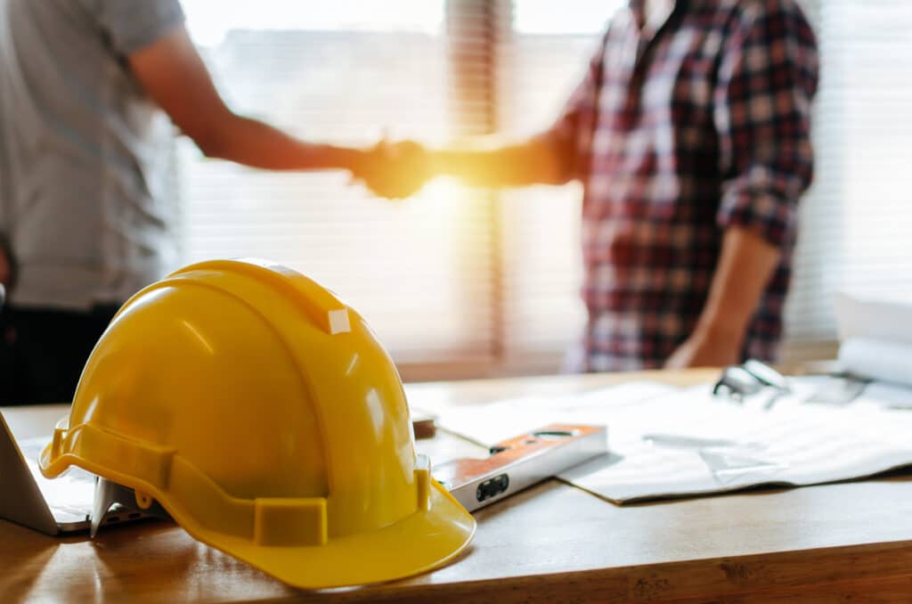 Two men shaking hands at a desk, both in hard hats, representing their partnership with Gulf Coast Mats for construction projects.